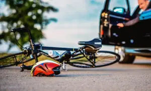 Wypadek na drodze. Rower leży na ziemi obok kasku. Kierowca wysiada z samochodu. Zastanów się, co grozi za potrącenie rowerzysty.