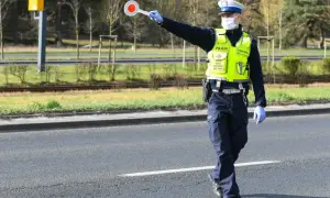 Policjant w kamizelce odblaskowej i masce zatrzymuje ruch, trzymając tarczę. Nie zatrzymanie się do kontroli grozi konsekwencjami.