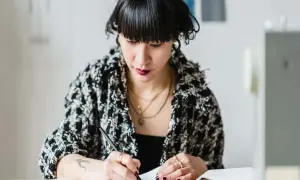 Kobieta w eleganckiej marynarce pochyla się nad dokumentami, pisząc. Rozważa umowę zlecenie i składki obowiązkowe.