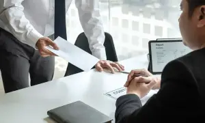 Przełożony wręcza pracownikowi dokumenty, być może umowa na czas określony. Na stole leży teczka i tablet z raportem.