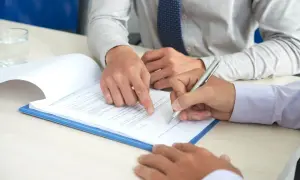 Dwóch mężczyzn przy stole, jeden wskazuje na dokument, drugi podpisuje. Może to być wezwanie jako świadek w sprawie art. 286 § 1 kk.