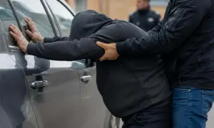 Mężczyzna w kapturze opiera się o samochód, gdy drugi mężczyzna go obezwładnia. W tle widać policjanta. Zastanawiamy się, ile grozi za takie zatrzymanie.
