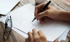 Ręka pisze długopisem na dokumencie, który może być wzorem pełnomocnictwa w sprawie spadkowej. Obok leżą okulary.