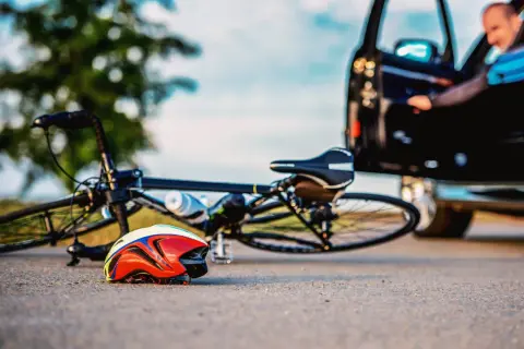 Wypadek na drodze. Rower leży na ziemi obok kasku. Kierowca wysiada z samochodu. Zastanów się, co grozi za potrącenie rowerzysty.