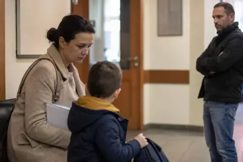 Rodzina w sądzie, matka z synem siedzą, ojciec stoi z boku. Sytuacja sugeruje wniosek o ustalenie kontaktów z dzieckiem.