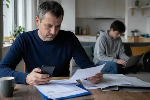 Ojciec z telefonem i dokumentami analizuje wydatki, podczas gdy syn pracuje przy laptopie. Myśli o obowiązek alimentacyjny do 26 roku życia.