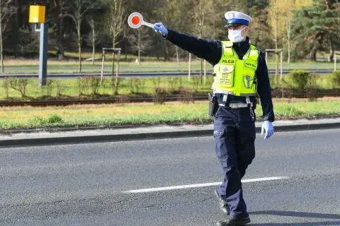Policjant w kamizelce odblaskowej i masce zatrzymuje ruch, trzymając tarczę. Nie zatrzymanie się do kontroli grozi konsekwencjami.