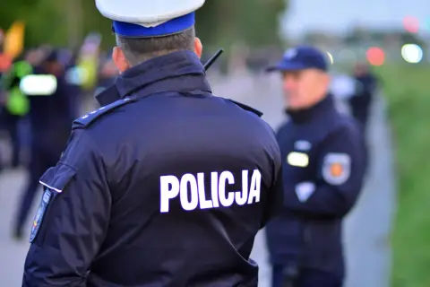 Policjant w mundurze z napisem "POLICJA" na plecach. Czy policjant może mnie przeszukać na ulicy? W tle inni funkcjonariusze i niewyraźne sylwetki.