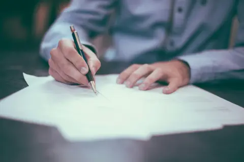 Dłoń pisze na dokumencie, co może być związane z fałszowaniem dokumentów i wyrokami sądowymi.