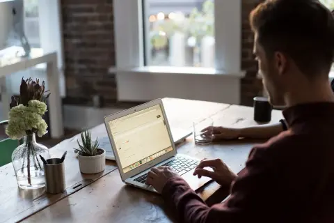 Mężczyzna pracuje na laptopie, nawet w sobotę. W tle widać okno i kwiaty.