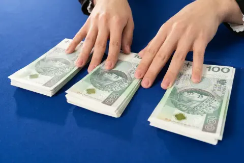 Dłonie nad stosem banknotów 100 zł. Symbolizuje to darowiznę między małżonkami, wsparcie finansowe lub oszczędności.