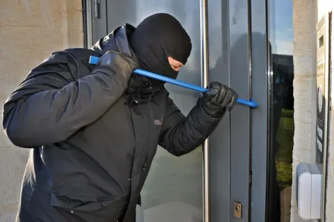 Mężczyzna w kominiarce i rękawiczkach próbuje wyważyć drzwi łomem. To próba kradzieży z włamaniem.