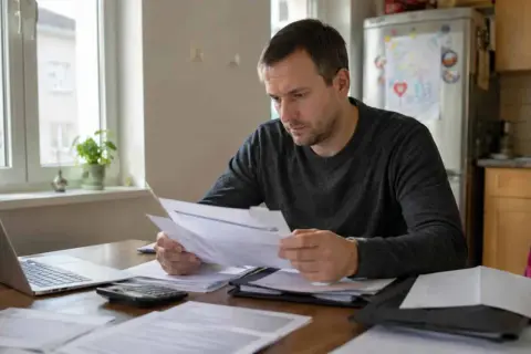 Mężczyzna analizuje dokumenty, przygotowując wniosek o obniżenie alimentów. Na stole laptop, kalkulator i sterta papierów.