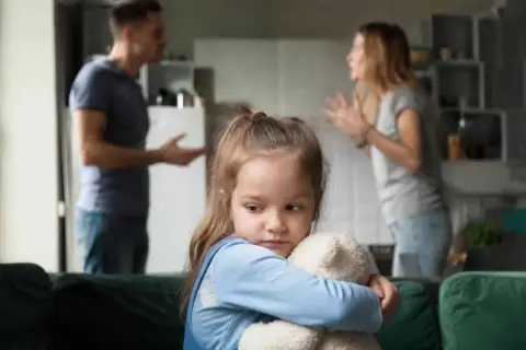 Smutna dziewczynka tuli misia, podczas gdy rodzice kłócą się w tle. Temat: pozbawienie praw rodzicielskich a alimenty.