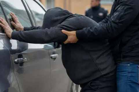 Mężczyzna w kapturze opiera się o samochód, gdy drugi mężczyzna go obezwładnia. W tle widać policjanta. Zastanawiamy się, ile grozi za takie zatrzymanie.