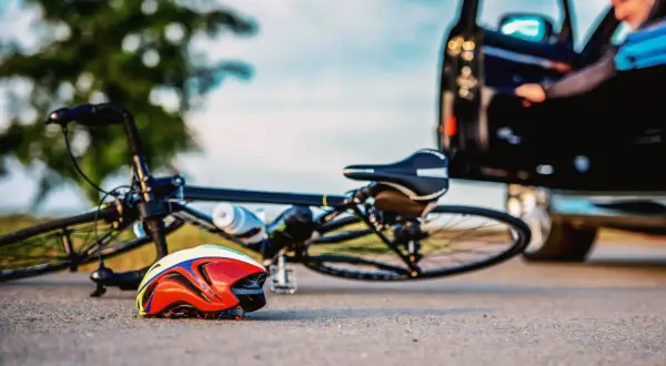 Wypadek na drodze. Rower leży na ziemi obok kasku. Kierowca wysiada z samochodu. Zastanów się, co grozi za potrącenie rowerzysty.