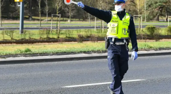Policjant w kamizelce odblaskowej i masce zatrzymuje ruch, trzymając tarczę. Nie zatrzymanie się do kontroli grozi konsekwencjami.