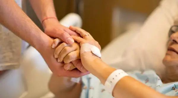 Dłoń opiekuna obejmuje dłoń osoby starszej w łóżku. Po śmierci osoby ubezwłasnowolnionej opiekun prawny ma obowiązki.