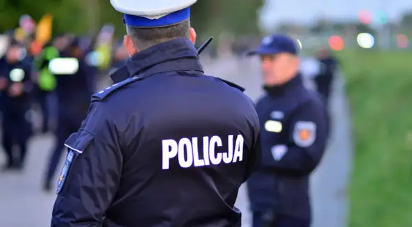 Policjant w mundurze z napisem "POLICJA" na plecach. Czy policjant może mnie przeszukać na ulicy? W tle inni funkcjonariusze i niewyraźne sylwetki.