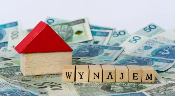 Drewniany domek i kostki z napisem "WYNAJEM" na tle polskich banknotów. Zastanawiasz się nad najmem okazjonalnym i podatkiem?
