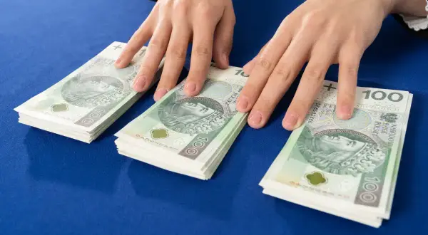Dłonie nad stosem banknotów 100 zł. Symbolizuje to darowiznę między małżonkami, wsparcie finansowe lub oszczędności.