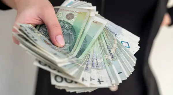 Dłoń trzyma plik banknotów 100 zł. Może to symbolizować zwrot kosztów za czynsz od spadkobierców.