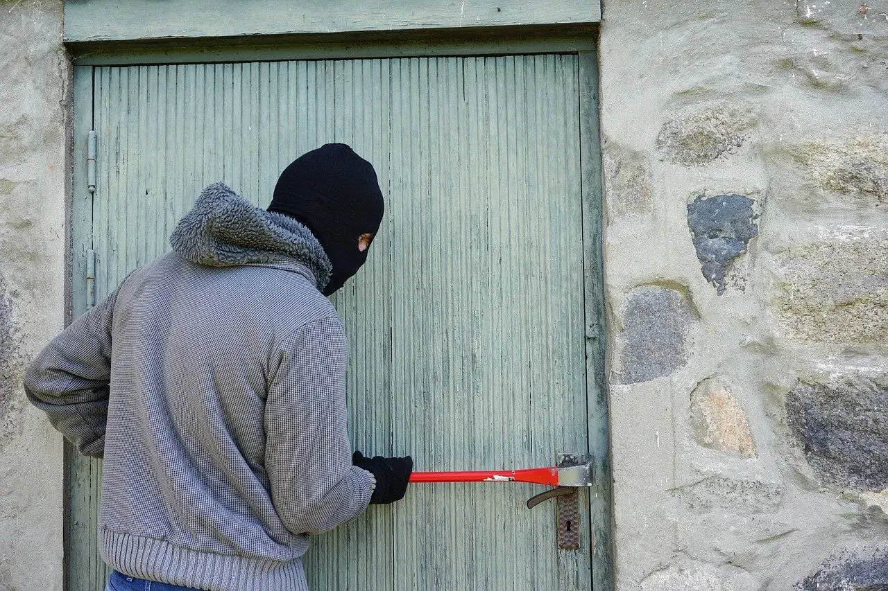 Osoba w masce próbuje wyważyć drzwi łomem. To próba kradzieży z włamaniem.