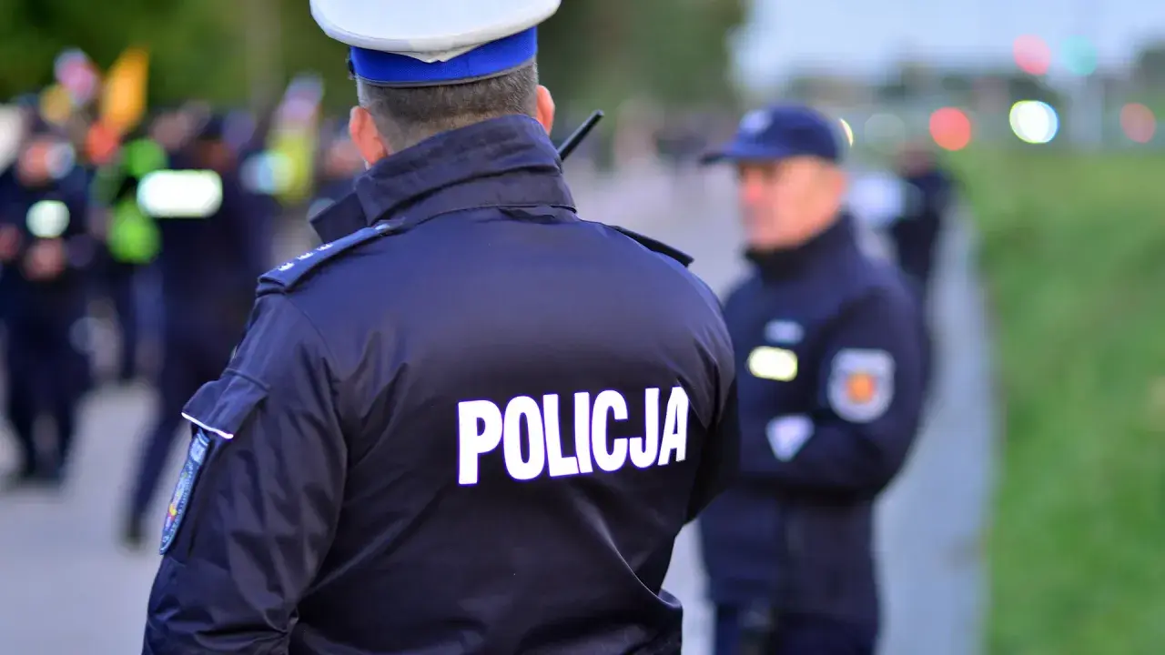 Policjant w mundurze z napisem "POLICJA" na plecach. Czy policjant może mnie przeszukać na ulicy? W tle inni funkcjonariusze i niewyraźne sylwetki.
