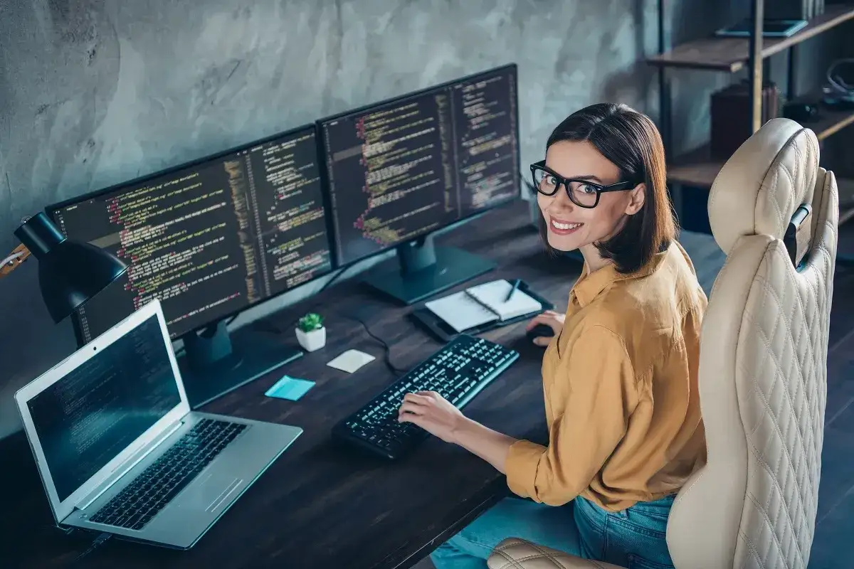 Uśmiechnięta programistka w okularach pracuje przy wielu monitorach. Jej zadaniowy czas pracy pozwala na elastyczność i efektywność.
