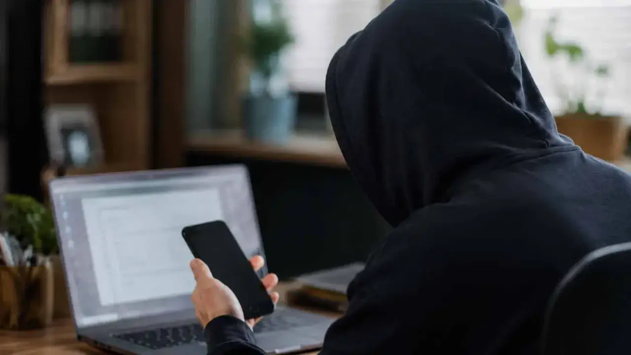 Osoba w kapturze przy laptopie i telefonie. Czy bank odda skradzione pieniądze z konta?