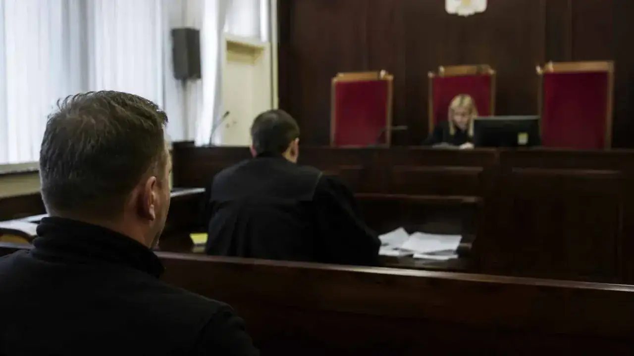 Mężczyzna w czerni, obrońca w sądzie, czeka na rozpoczęcie rozprawy. Sędzia i ławnicy siedzą za stołem.