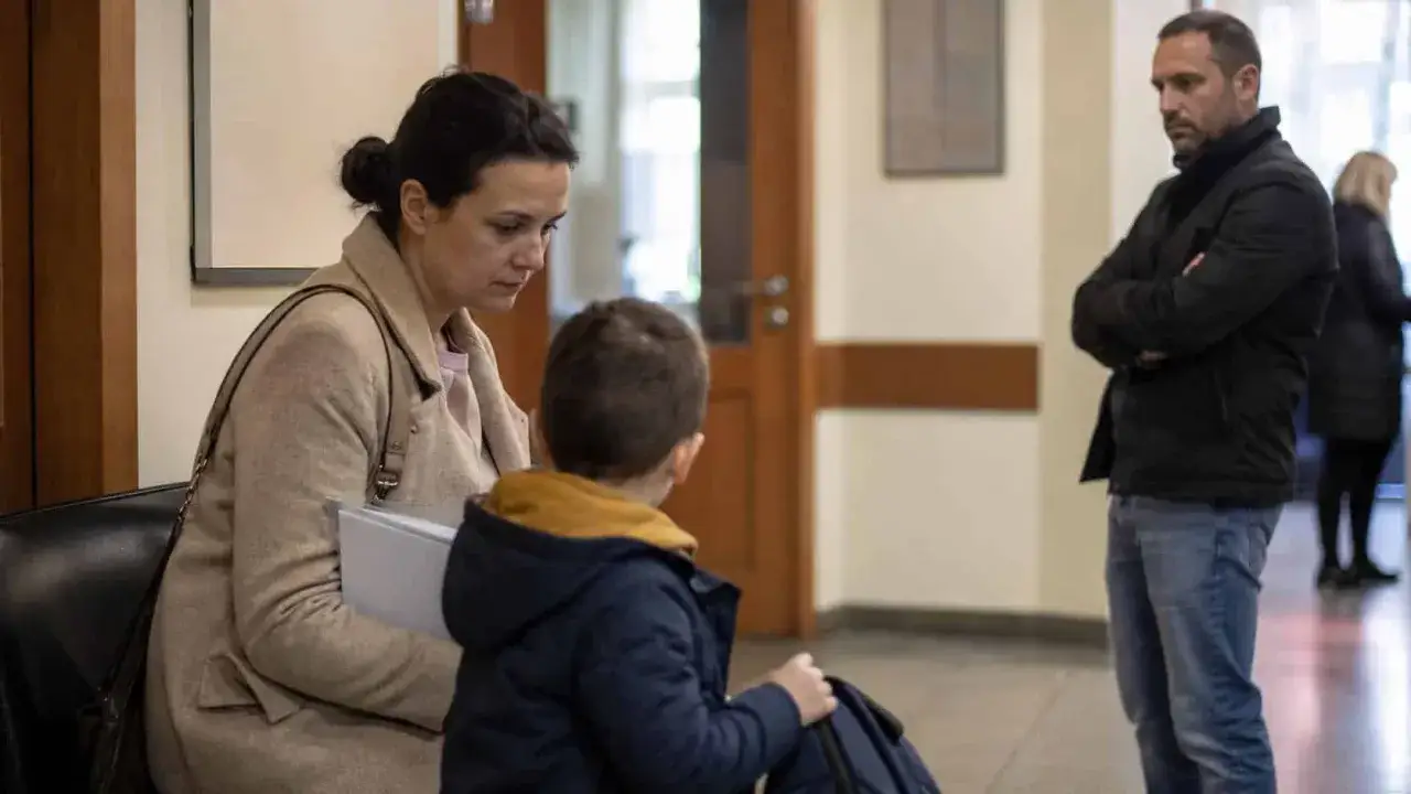 Ojciec z założonymi rękami czeka, podczas gdy matka z synem siedzą. Sytuacja może dotyczyć praw ojca płacącego alimenty.