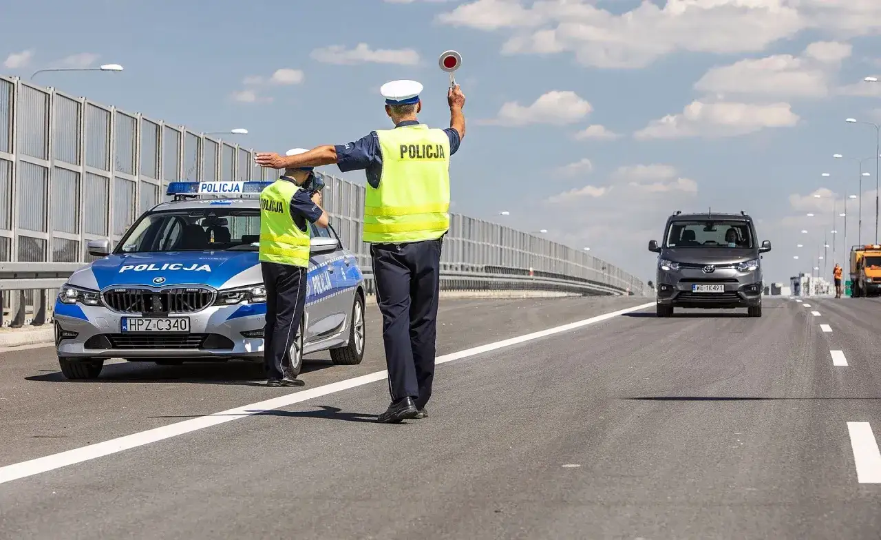 Policjanci kierują ruchem na drodze. Jeden z nich podnosi znak stop, sygnalizując kierowcy vana, że nie może kontynuować jazdy.