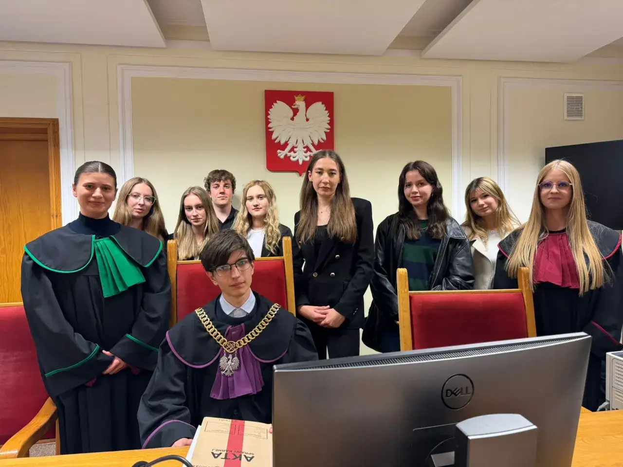 Grupa młodych ludzi w togach prawniczych, przygotowująca się do symulacji rozprawy. Jeden z nich trzyma akta sprawy karnej, sugerując możliwość umorzenia postępowania karnego.