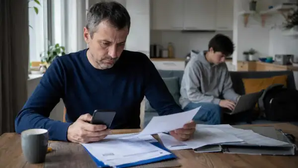 Ojciec z telefonem i dokumentami analizuje wydatki, podczas gdy syn pracuje przy laptopie. Myśli o obowiązek alimentacyjny do 26 roku życia.