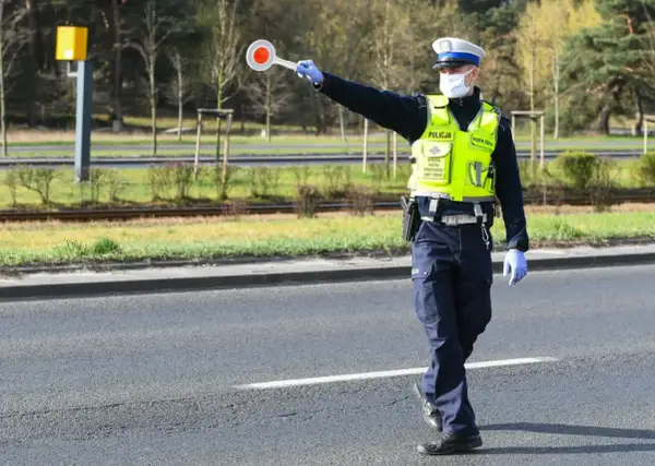 Policjant w kamizelce odblaskowej i masce zatrzymuje ruch, trzymając tarczę. Nie zatrzymanie się do kontroli grozi konsekwencjami.