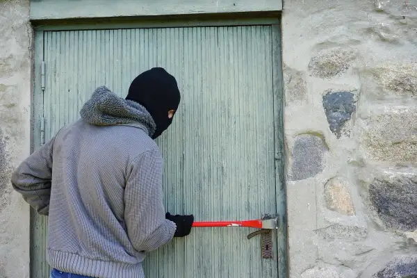 Osoba w masce próbuje wyważyć drzwi łomem. To próba kradzieży z włamaniem.