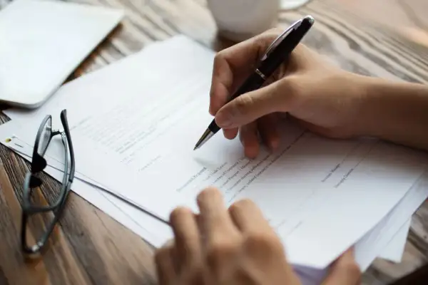 Dłoń pisze na dokumencie, który może być wzorem czynnego żalu. Obok leżą okulary i tablet.